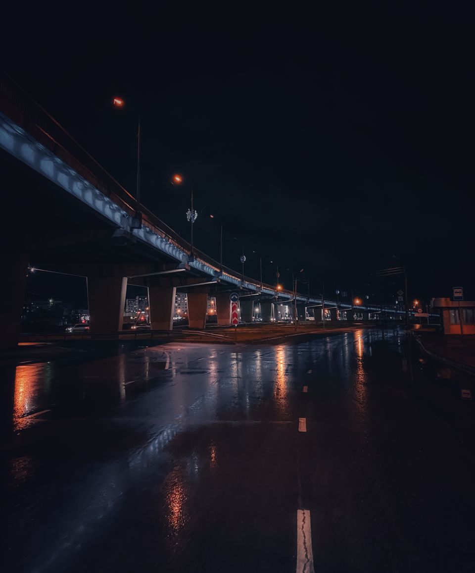 Wet asphalt at night under a highway overpass, amber street lamps reflecting in the standing water